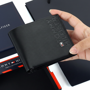 Hand holding a Tommy Hilfiger black smooth leather wallet featuring a large embossed vertical logo and the iconic small enamel flag
