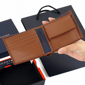 Handheld view of the open Tommy Hilfiger brown leather wallet alongside its branded gift box and luxury shopping bag