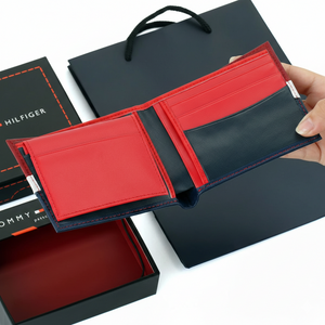 Hand holding open a Tommy Hilfiger men's color-block wallet showing a vibrant interior with red card slots on the left and a navy blue compartment on the right