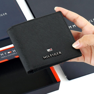 Hand holding a Tommy Hilfiger men's black leather bifold wallet with a small enamel flag logo plate on the corner, presented above its original navy blue gift box.