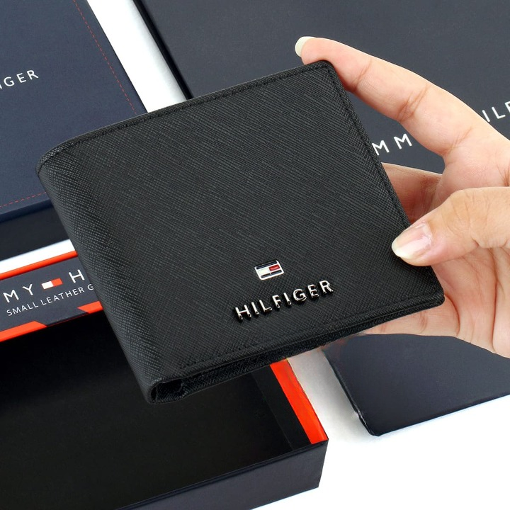 Hand holding a Tommy Hilfiger men's black leather bifold wallet with a small enamel flag logo plate on the corner, presented above its original navy blue gift box.