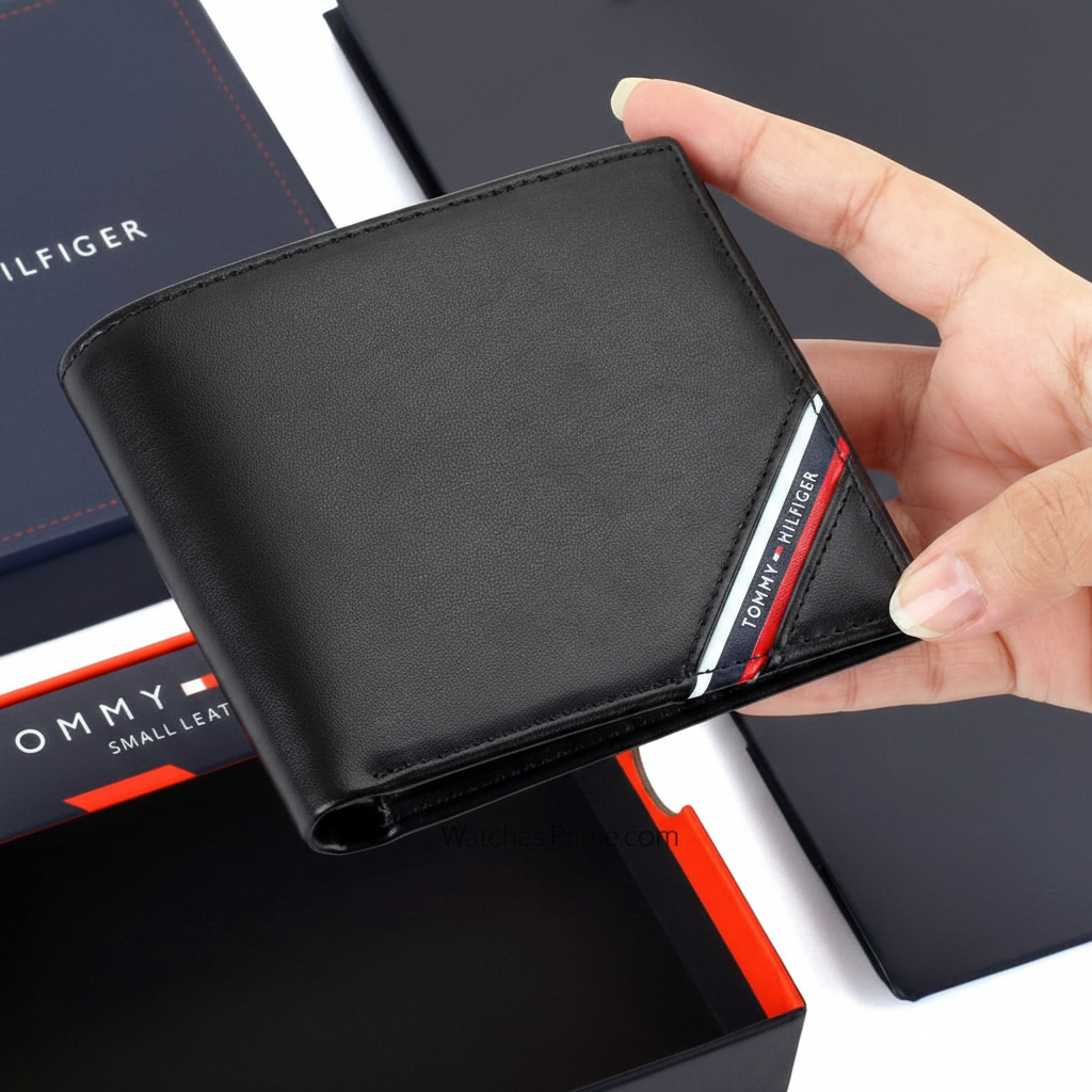 Handheld view of a Tommy Hilfiger men's black genuine leather wallet with the signature red, white, and blue flag detail, shown with its premium navy gift box