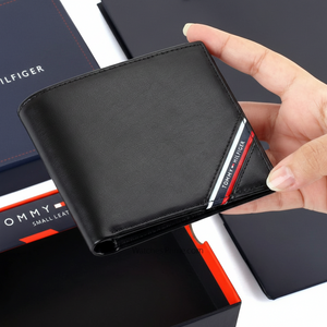 Handheld view of a Tommy Hilfiger men's black genuine leather wallet with the signature red, white, and blue flag detail, shown with its premium navy gift box