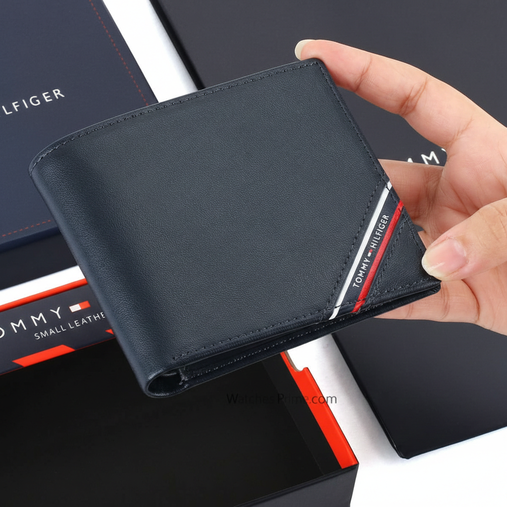 Hand holding a Tommy Hilfiger men's navy blue genuine leather wallet featuring a diagonal tricolor signature stripe and white brand lettering