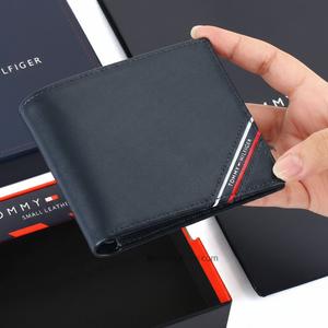 Hand holding a Tommy Hilfiger men's navy blue genuine leather wallet featuring a diagonal tricolor signature stripe and white brand lettering