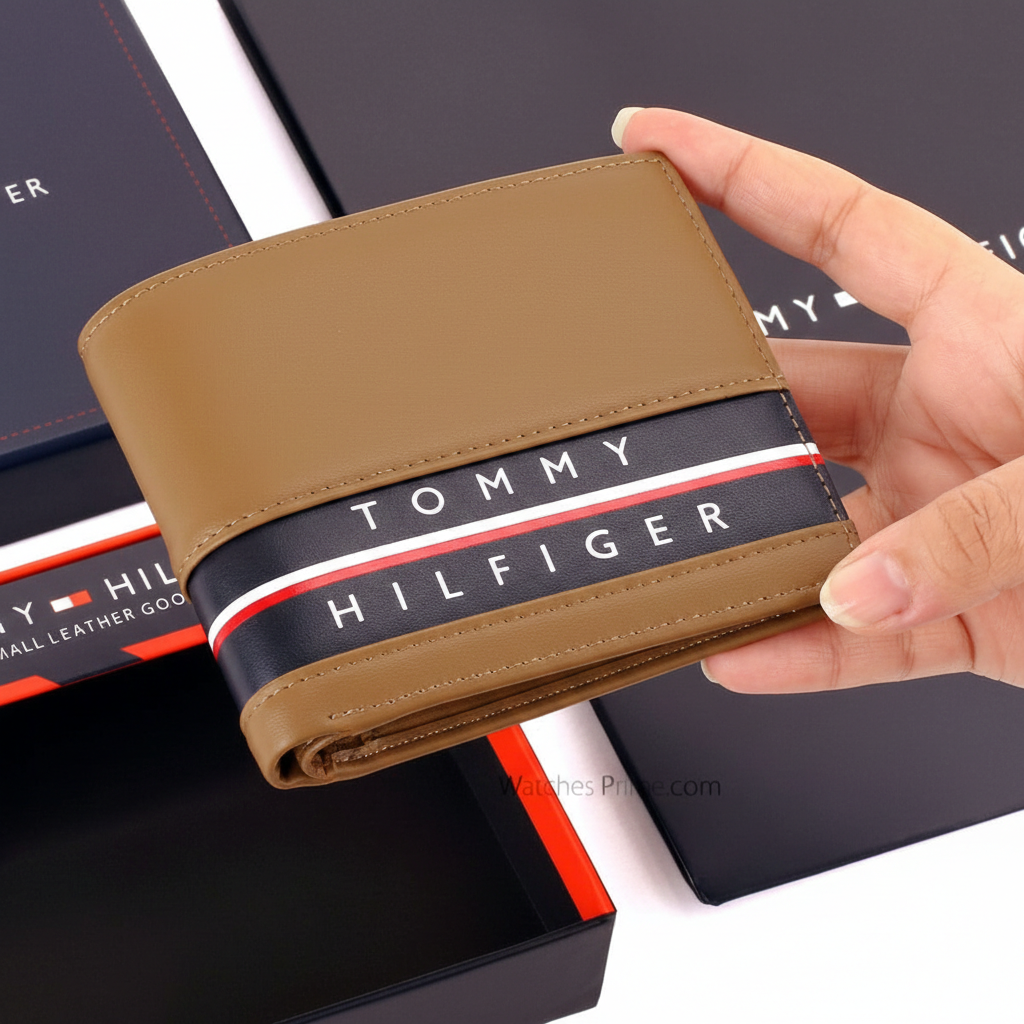 Handheld view of a Tommy Hilfiger men's tan leather wallet, showing its compact size and the vibrant red and white flag detail