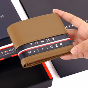 Handheld view of a Tommy Hilfiger men's tan leather wallet, showing its compact size and the vibrant red and white flag detail