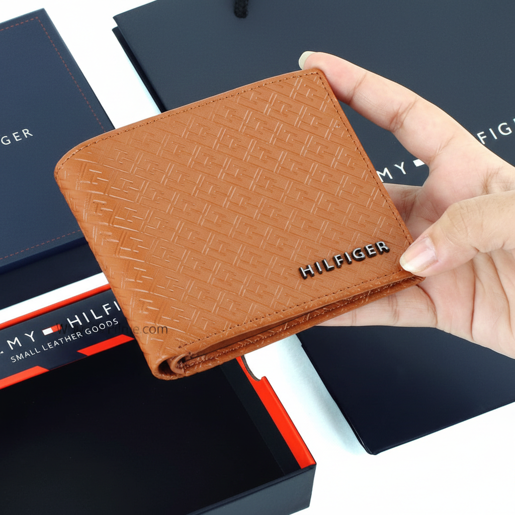 Hand holding a Tommy Hilfiger men's tan leather wallet featuring an all-over embossed Hilfiger monogram pattern and raised black metal logo