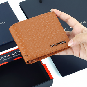 Hand holding a Tommy Hilfiger men's tan leather wallet featuring an all-over embossed Hilfiger monogram pattern and raised black metal logo